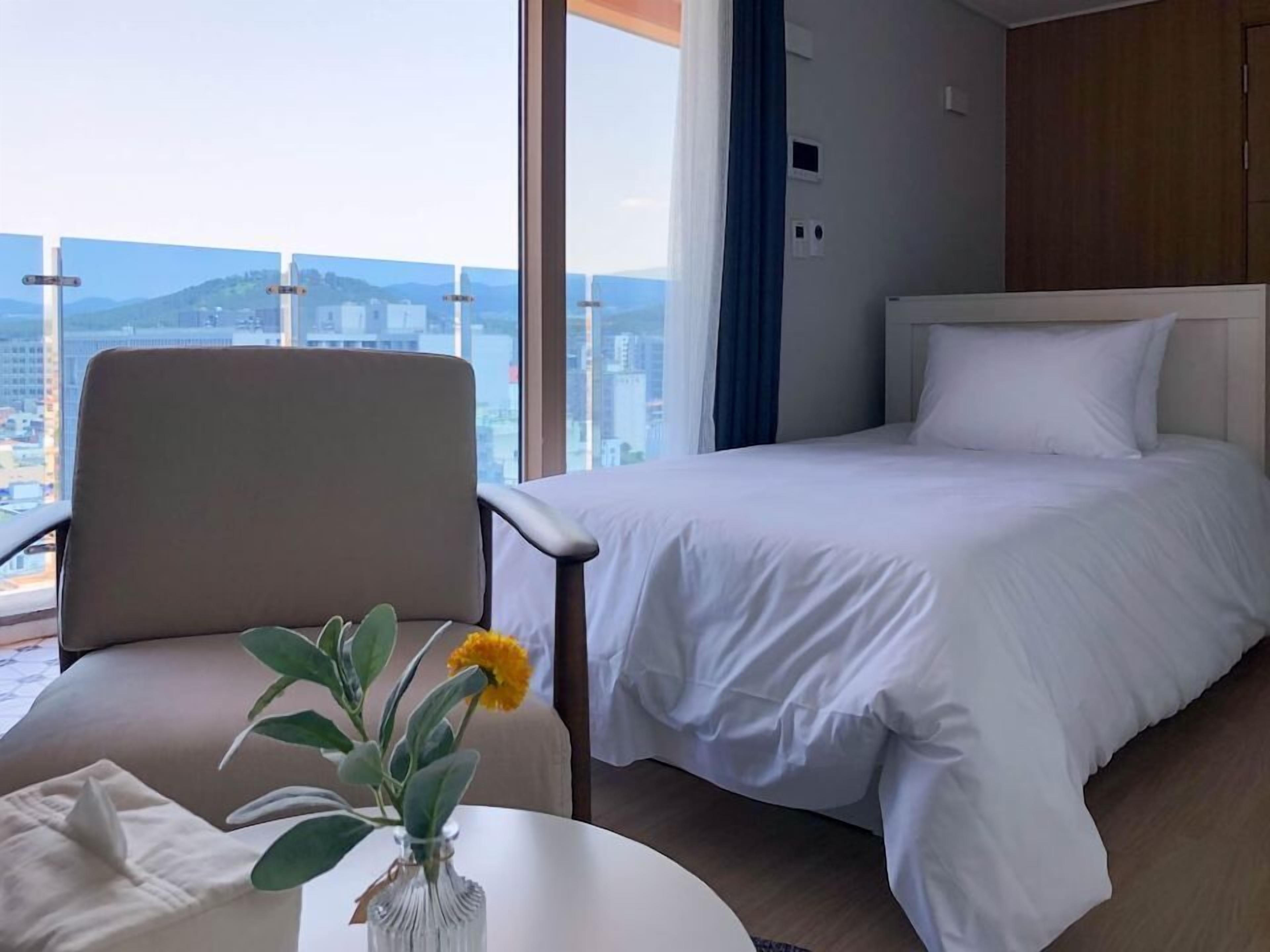 A hotel room with a bed, a chair, a small table with a vase of flowers, and a view of a cityscape through large windows.