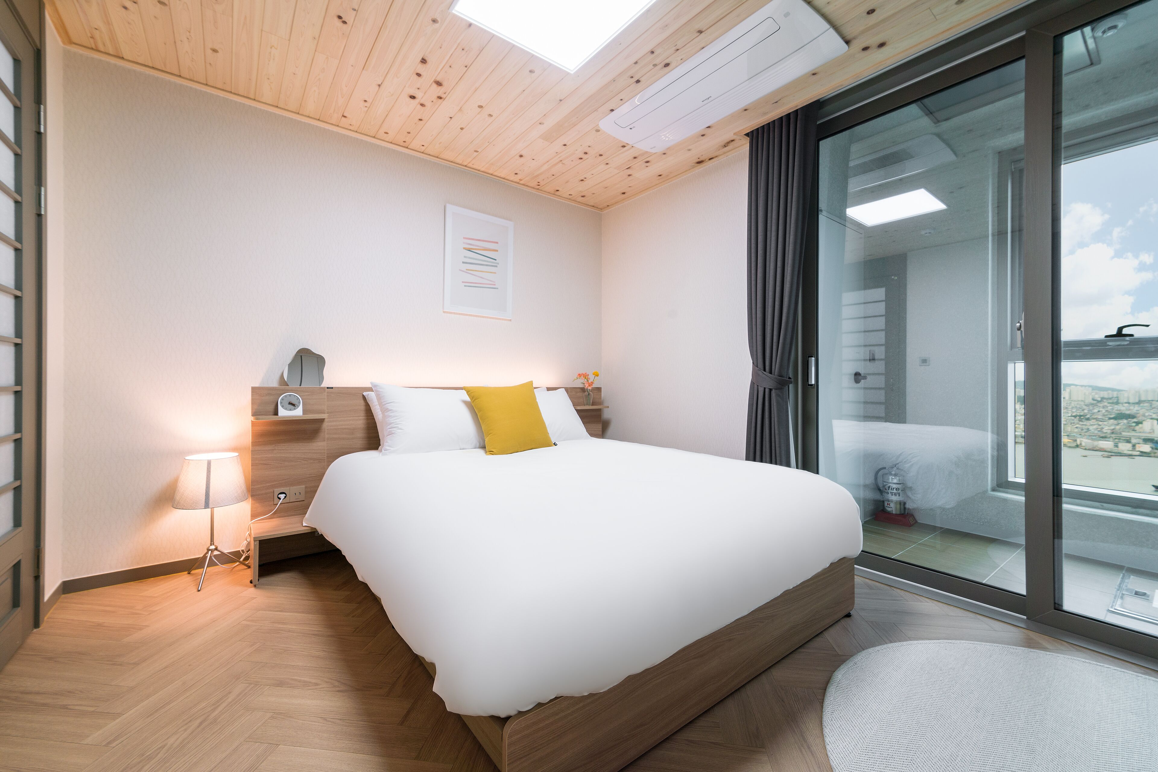 A modern bedroom with a wooden ceiling, a large bed, a bedside table, a lamp, and a view of the city through a sliding glass door.