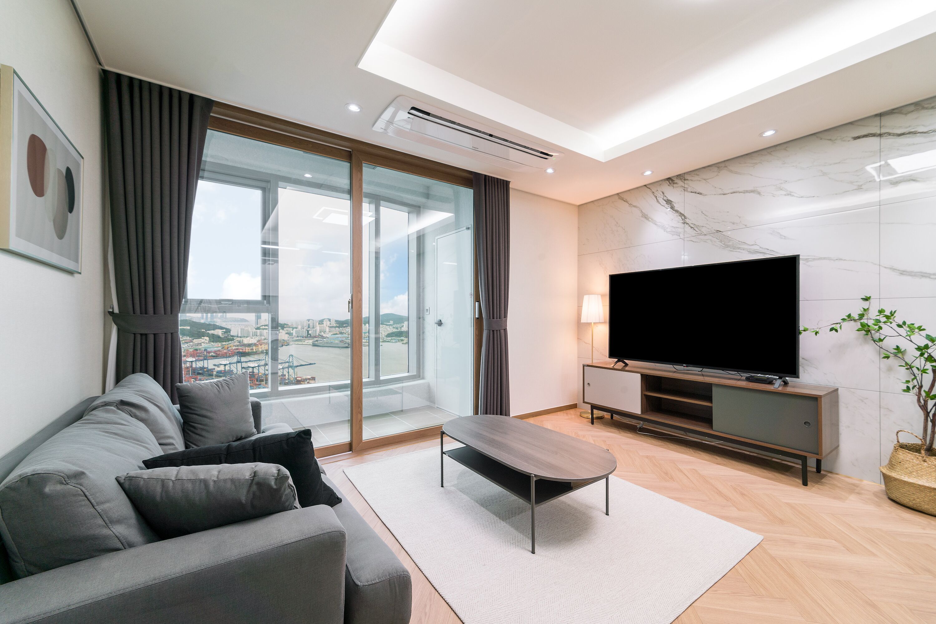 Un soggiorno moderno con un divano grigio, un tavolino da caffè, una TV a schermo piatto e un'ampia finestra vista sulla città.