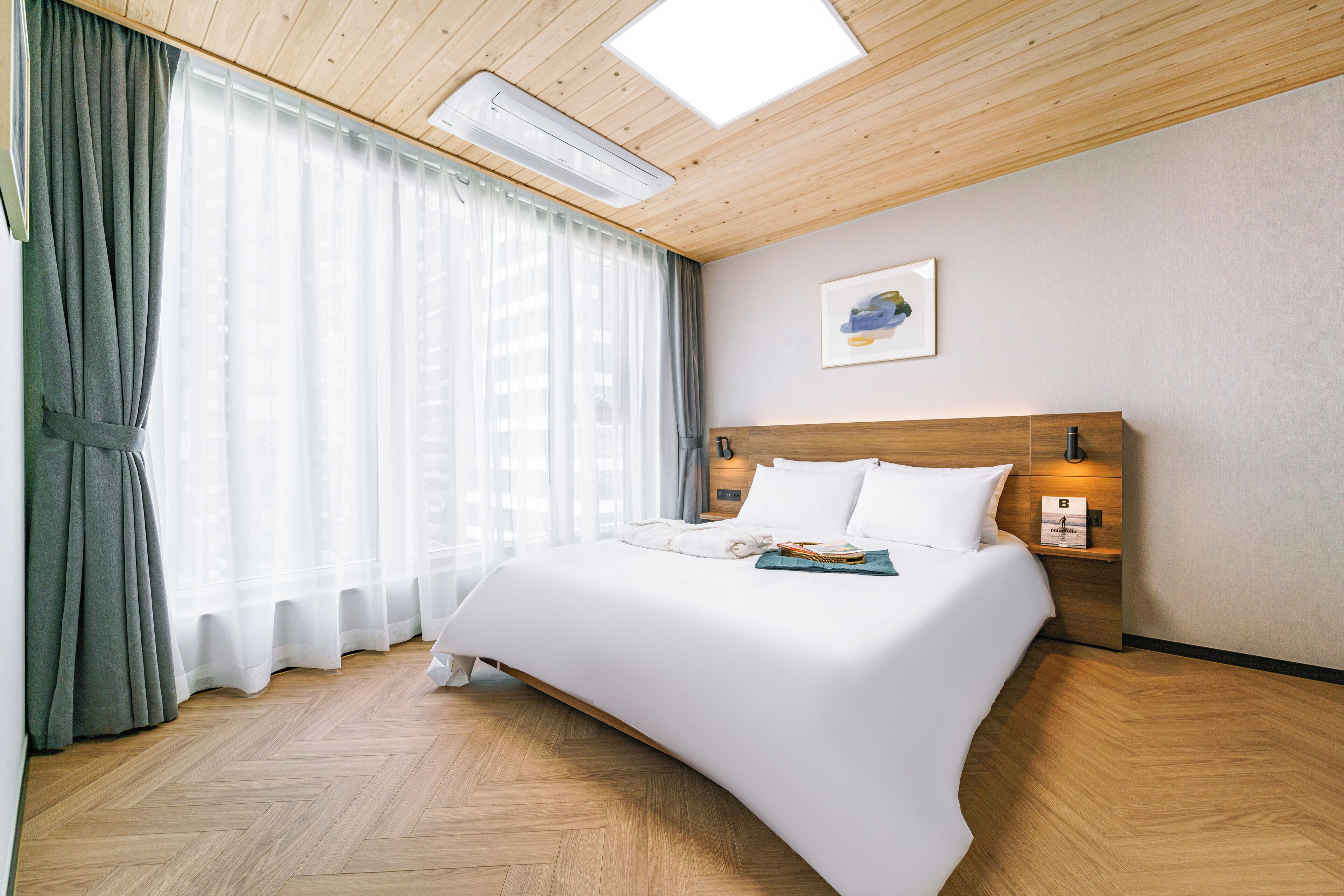 A hotel room with a wooden ceiling, a large bed with white bedding, a bedside table with a lamp, and a window with sheer curtains.