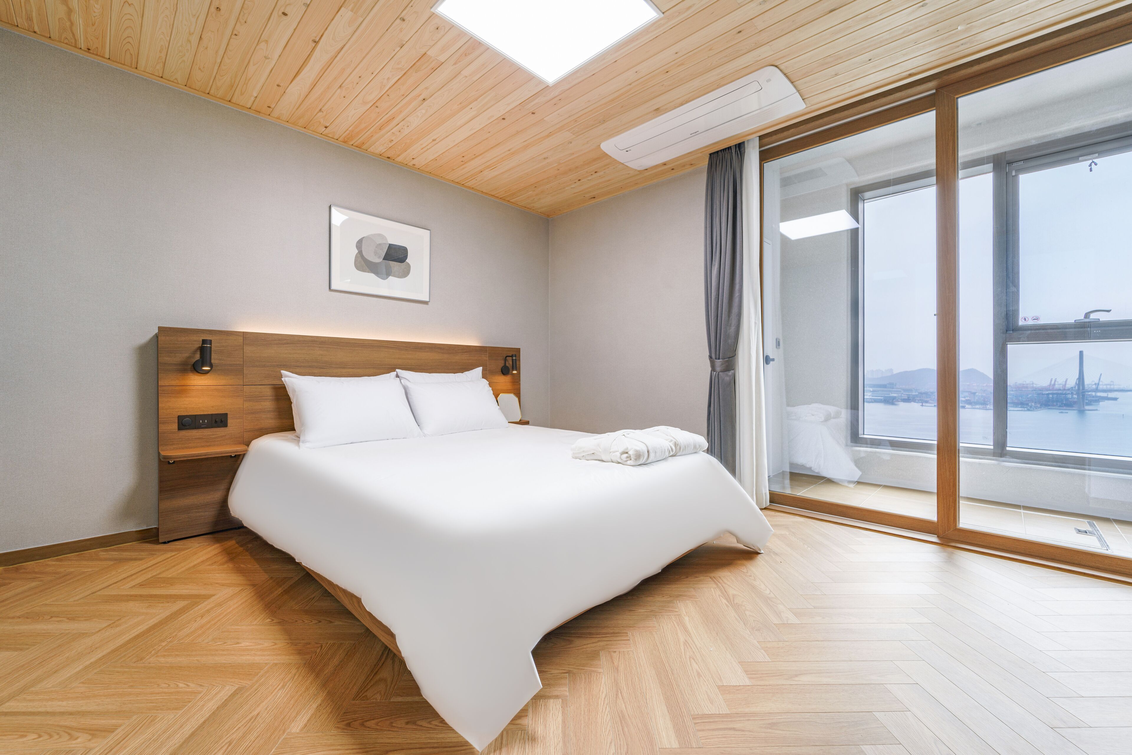 A modern hotel room with a wooden ceiling, a large bed, a view of a cityscape, and a sliding glass door.