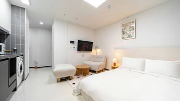 A modern hotel room with a large bed, a seating area with a small table, a TV mounted on the wall, and a washing machine in the corner.