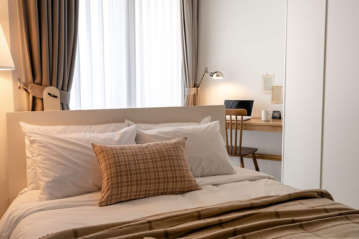 A neatly made bed with white and plaid pillows, a wooden desk with a laptop and a lamp, and a window with curtains.