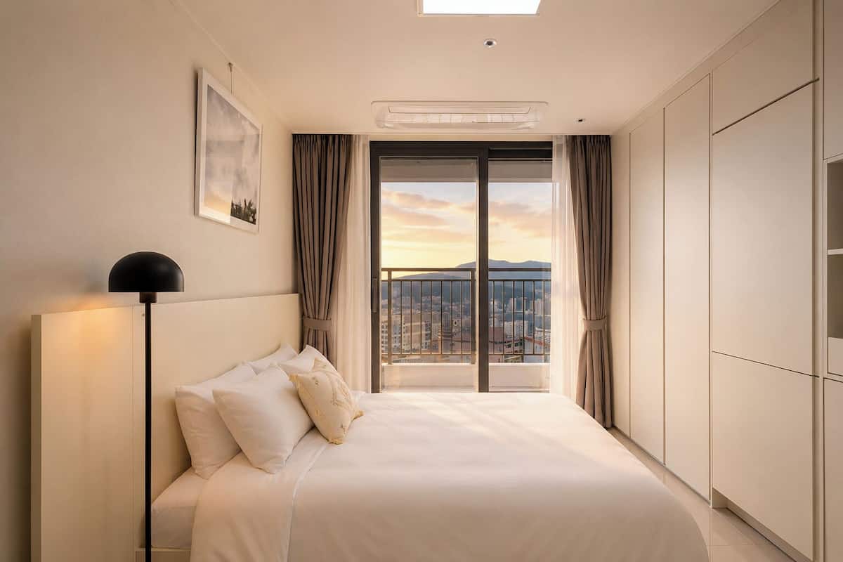 A hotel room with a bed, a floor lamp, a view of the city through a balcony, and a ceiling light.