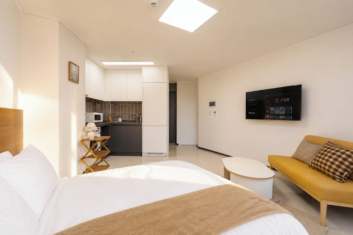 A modern hotel room with a bed, a sofa, a small table, a TV, and a kitchenette.