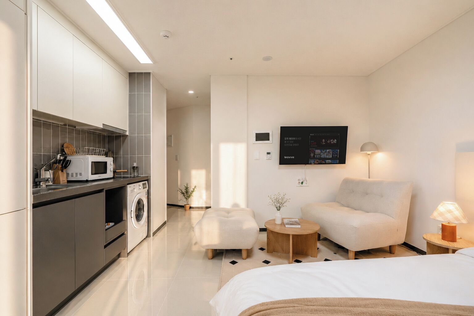 A modern hotel room with a kitchenette, a flat-screen TV, a sofa, a bed, and a small round table.