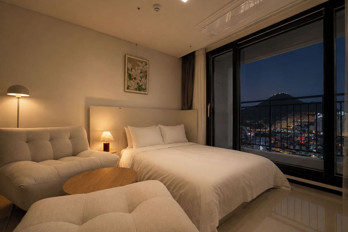 A modern hotel room with a large bed, a sofa, a small table, and a city view through a balcony.