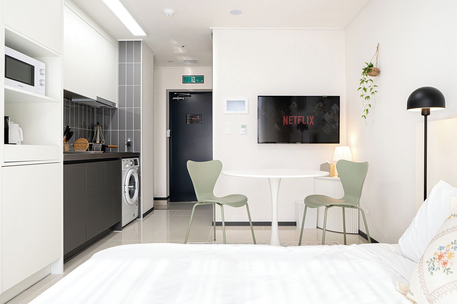Una habitació d'hotel moderna amb un llit, una cuineta petita, una zona de menjador amb dues cadires, un televisor i una làmpada de nit.
