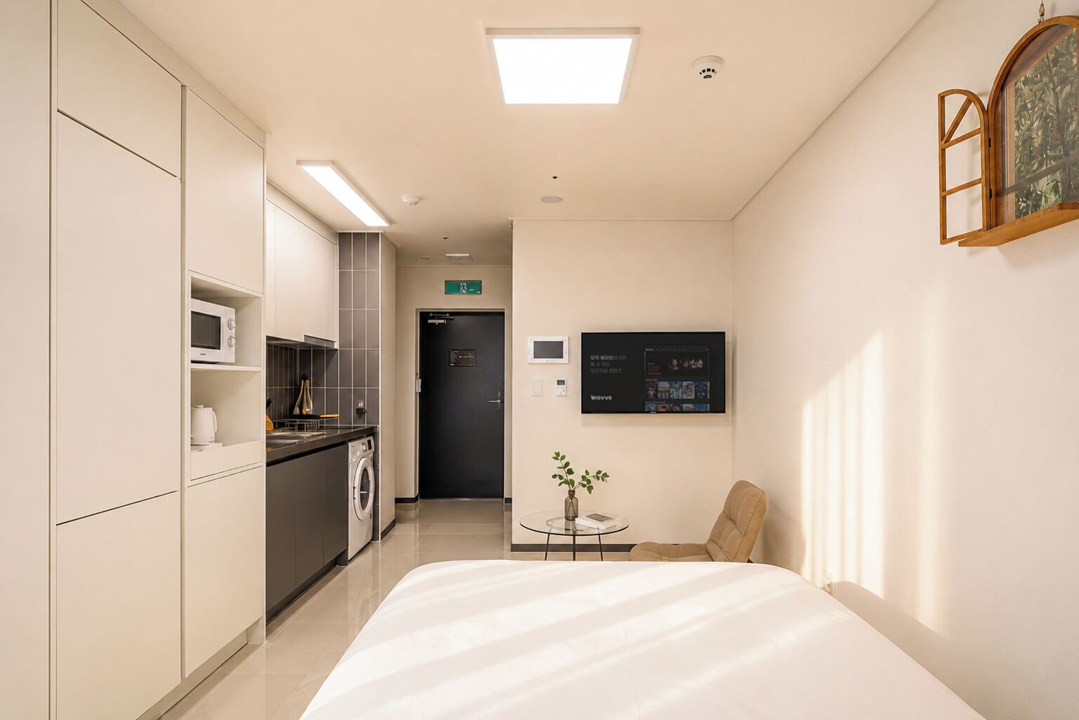 A hotel room with a bed, a small table with a vase, a TV on the wall, and a kitchenette with a microwave and washing machine.