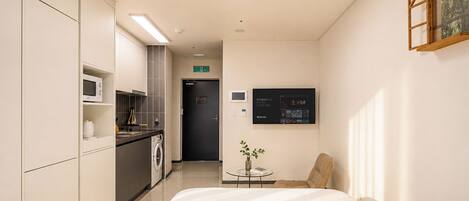 A hotel room with a bed, a small table with a vase, a TV on the wall, and a kitchenette with a microwave and washing machine.