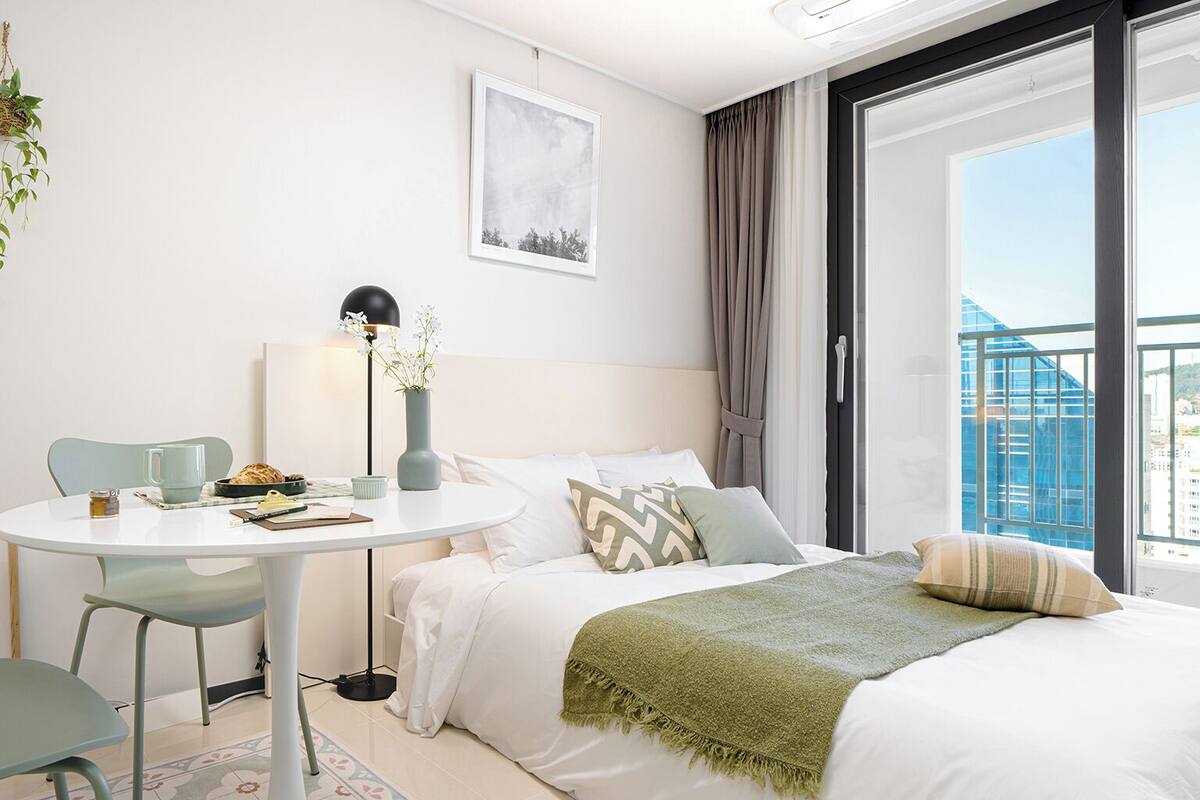 A modern hotel room with a bed, a round dining table, and a chair. There is a window with a view of a building and a balcony.