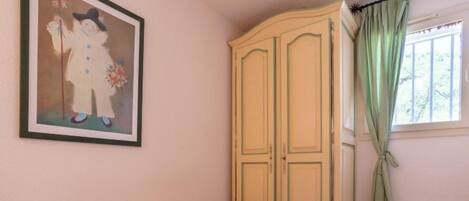 A bedroom with a floral bedspread, a framed picture on the wall, a wardrobe, and a window with curtains.