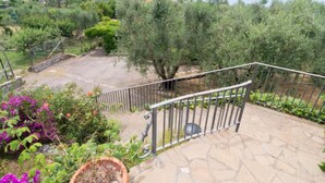 Une terrasse en pierre avec une rambarde en métal, entourée de verdure et d’oliviers, offrant une vue sur un paysage côtier.