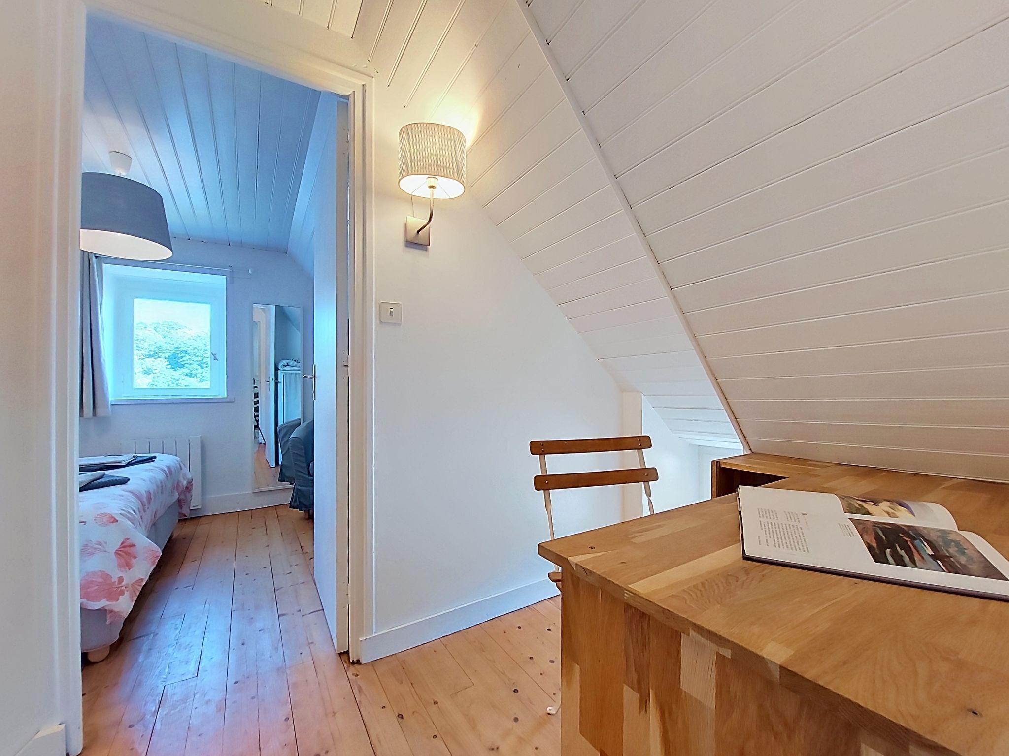 Ein Zimmer mit schrägen Wänden, einem Bett mit floralem Überwurf, einem Holztisch mit einer Zeitschrift und einem Fenster mit Blick auf Bäume.