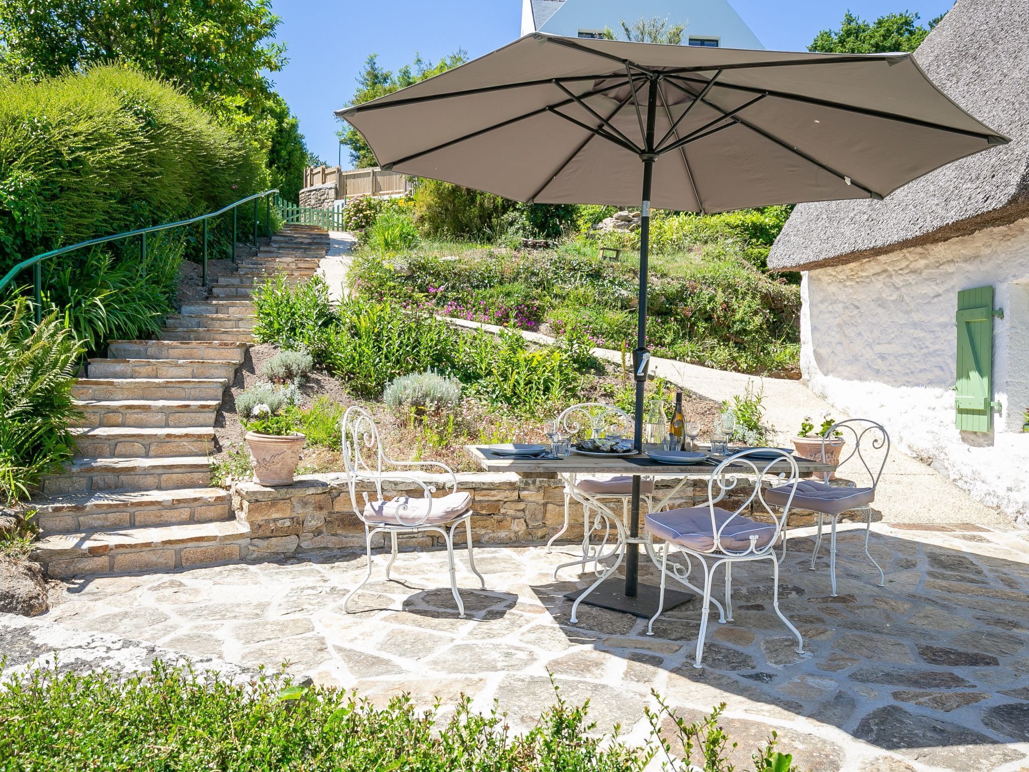 A stone patio with a table and chairs, a large umbrella, and a staircase leading up to a house.