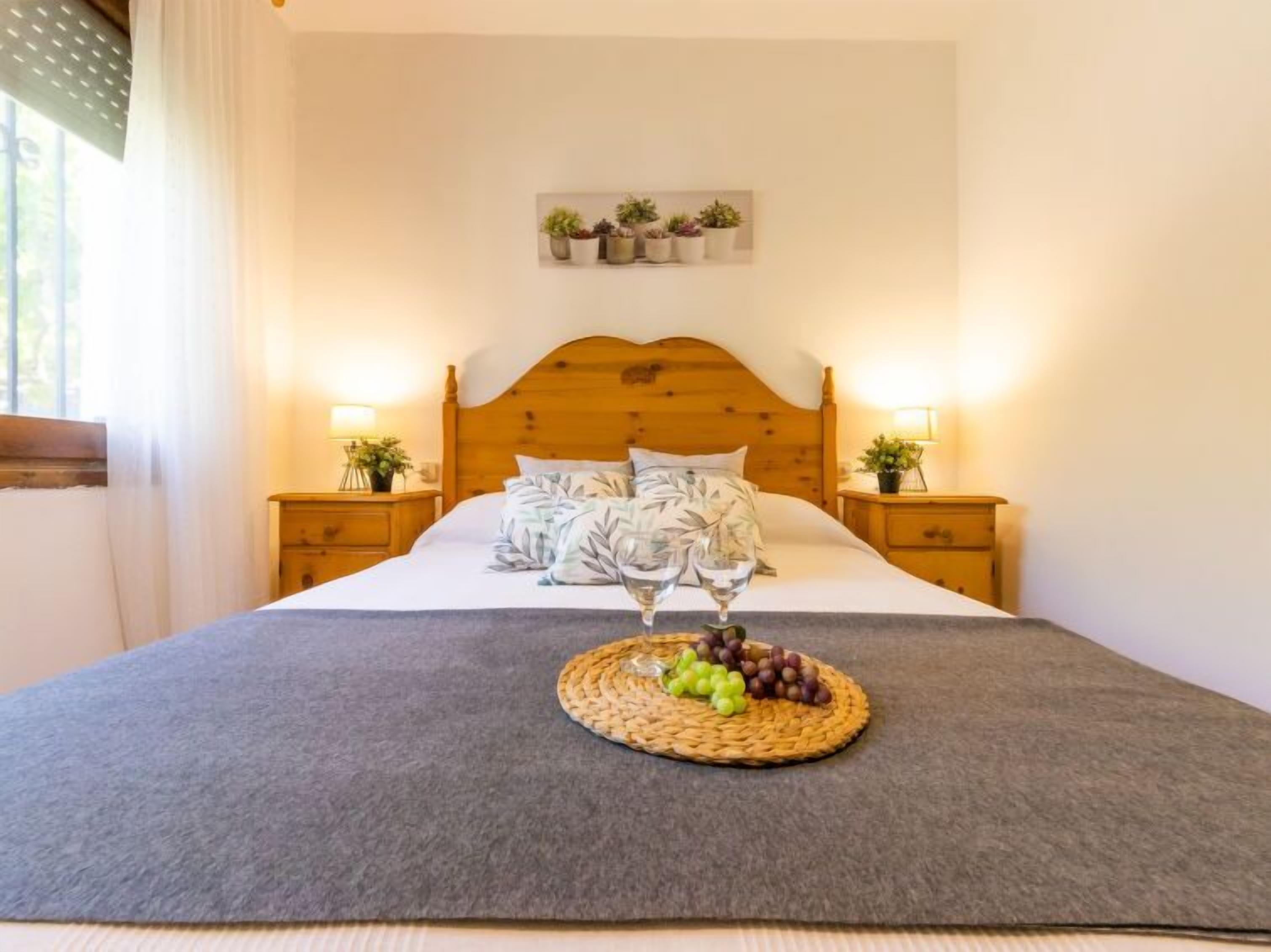 Une chambre avec une tête de lit en bois, deux tables de chevet avec des lampes, un lit recouvert d’une couverture grise, une étagère à plantes fixée au mur et un panier de fruits posé sur le lit.