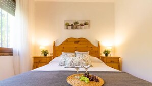 A bedroom with a wooden headboard, two bedside tables with lamps, a bed with a grey blanket, a wall-mounted plant shelf, and a fruit basket on the bed. - Vacation home Moru by Interhome (Deltebre)
