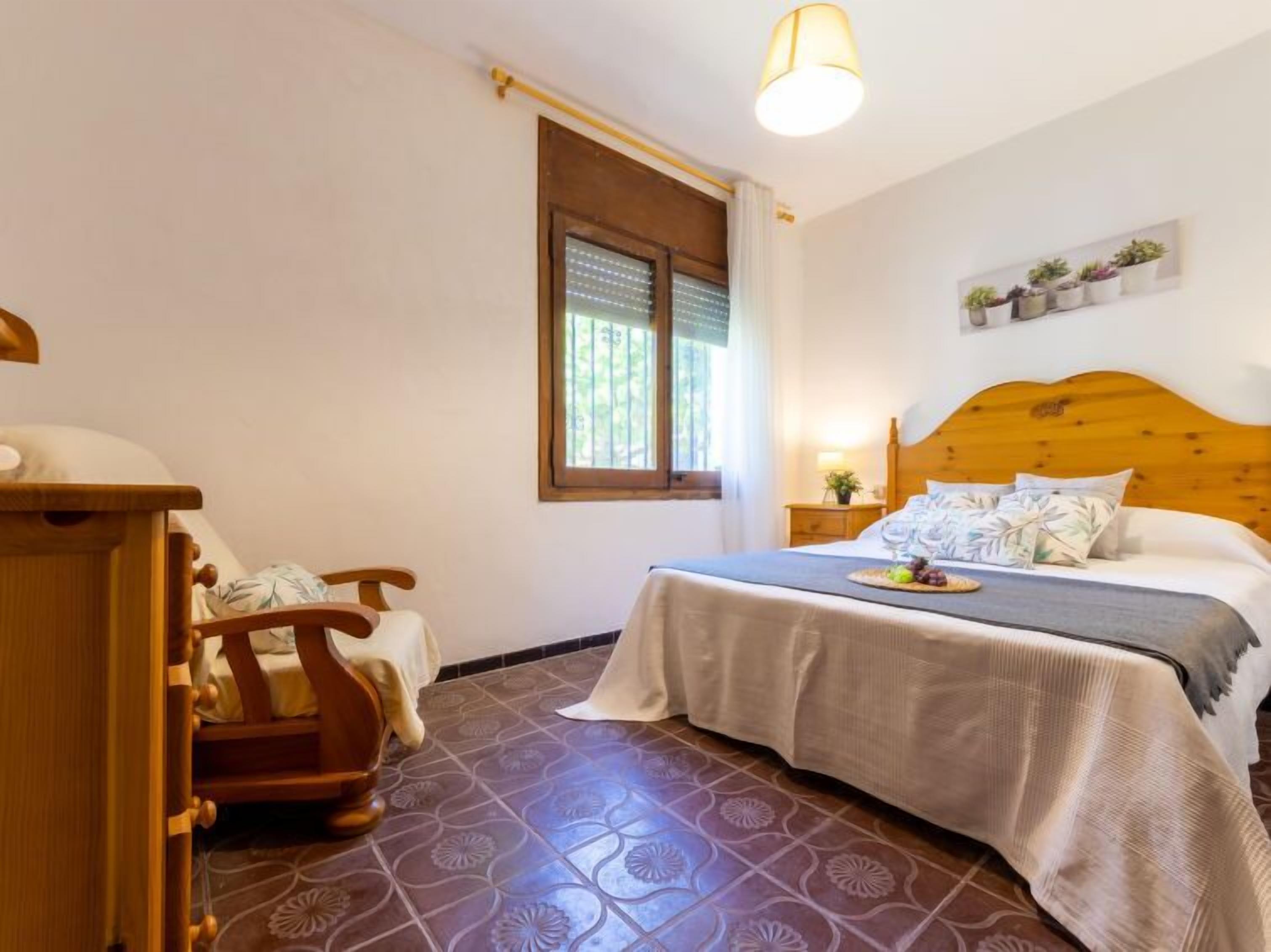 Une chambre avec une tête de lit en bois, un lit recouvert d’une couverture grise, une table de chevet avec une lampe, une fenêtre avec des stores et une étagère murale ornée de plantes.