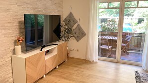 A modern living room with a flat-screen TV on a white console, a wooden floor, and a sliding glass door leading to an outdoor patio with a table and chairs. - JUPAS° | Red Rock Home - Balcony & Parking lot (Trier)