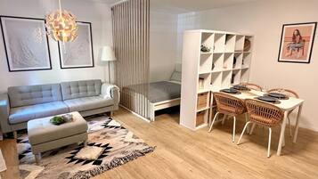 A modern living room with a sofa, a small bed, a dining area with wicker chairs, and a bookshelf.