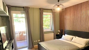 A bedroom with a bed, a dresser, a nightstand, a lamp, and a window with curtains. - JUPAS° | Red Rock Home - Balcony & Parking lot (Trier)
