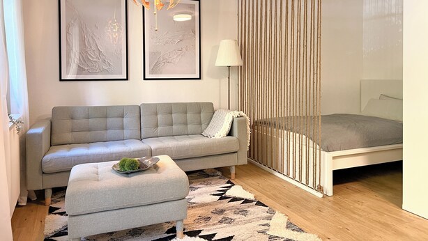 A modern bedroom with a grey sofa, a small ottoman, a bed with a white headboard, a hanging light fixture, and two framed abstract artworks on the wall. - JUPAS° | Red Rock Home - Balcony & Parking lot (Trier)