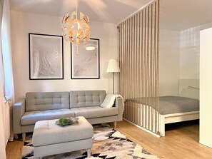 A modern bedroom with a grey sofa, a small ottoman, a bed with a white headboard, a hanging light fixture, and two framed abstract artworks on the wall. - JUPAS° | Red Rock Home - Balcony & Parking lot (Trier)