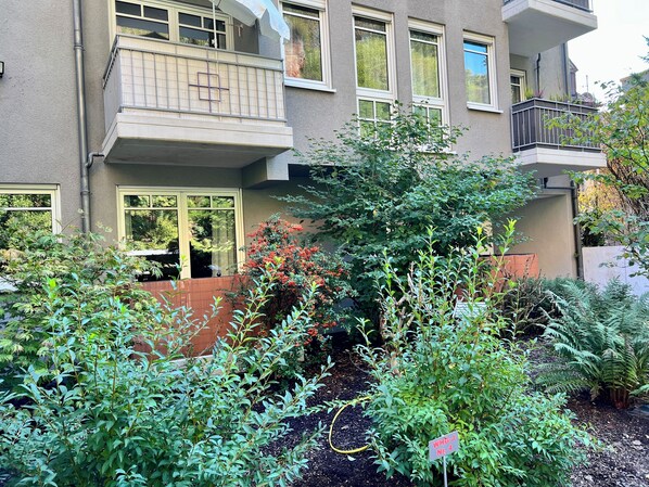 A residential building with a balcony, a garden with various plants, and a sign indicating a specific area. - JUPAS° | Red Rock Home - Balcony & Parking lot (Trier)