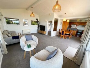 A spacious living area with a sofa, armchairs, a coffee table, and a dining table with chairs. There is a television and a kitchen area visible in the background.