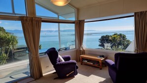 A spacious room with large windows offering a view of the sea and distant mountains. There are two purple armchairs and a small wooden table with books.