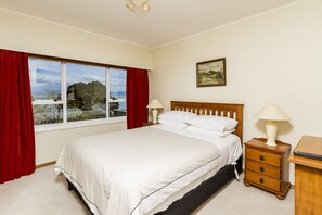 A bedroom with a large bed, a nightstand with a lamp, a wooden dresser, and a window with red curtains. - Kimberley House - 6-bedroom home in a fantastic Nelson waterfront location. (Nelson)