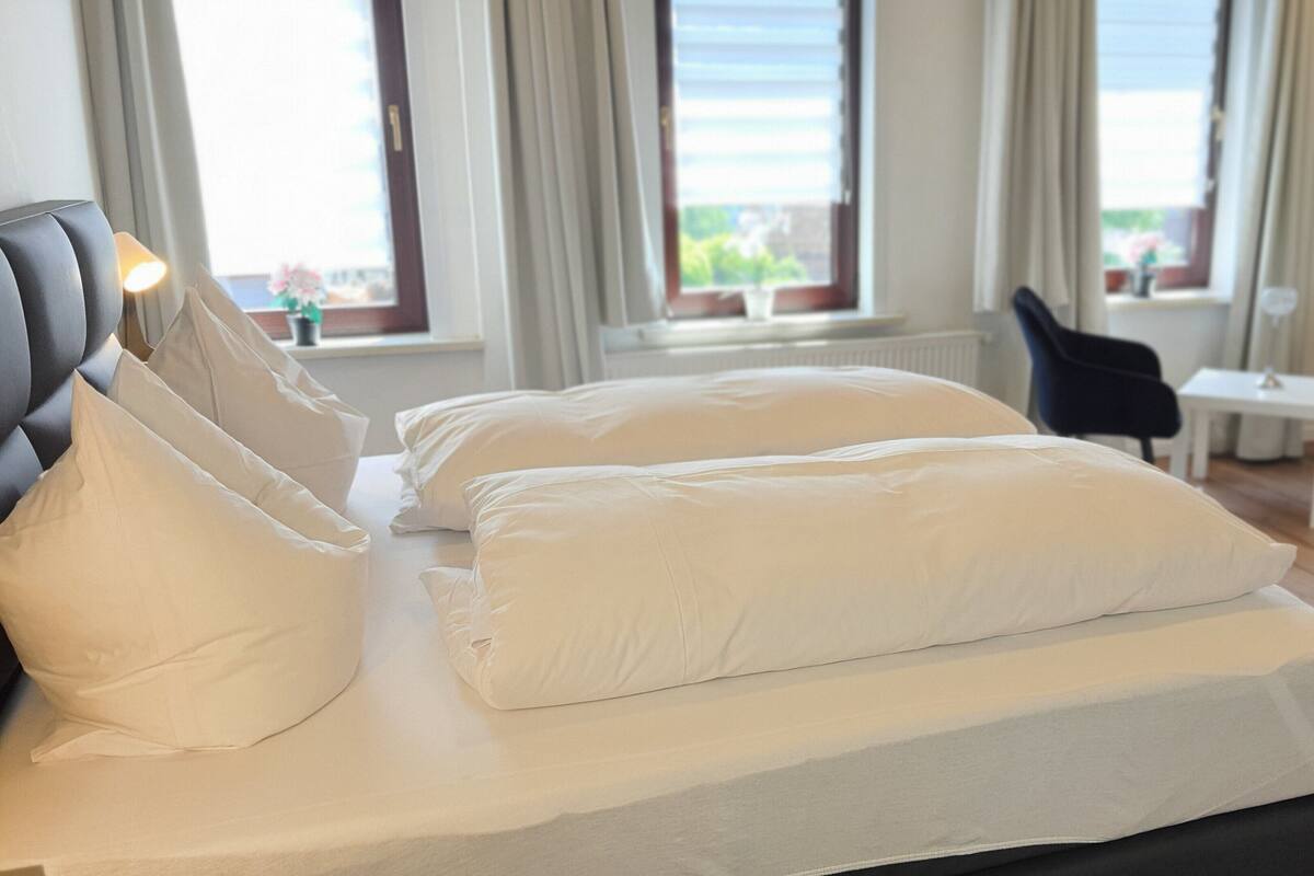 Un lit avec du linge de lit blanc et une tête de lit, une petite table et une chaise, dans une pièce dotée de grandes fenêtres et de rideaux.
