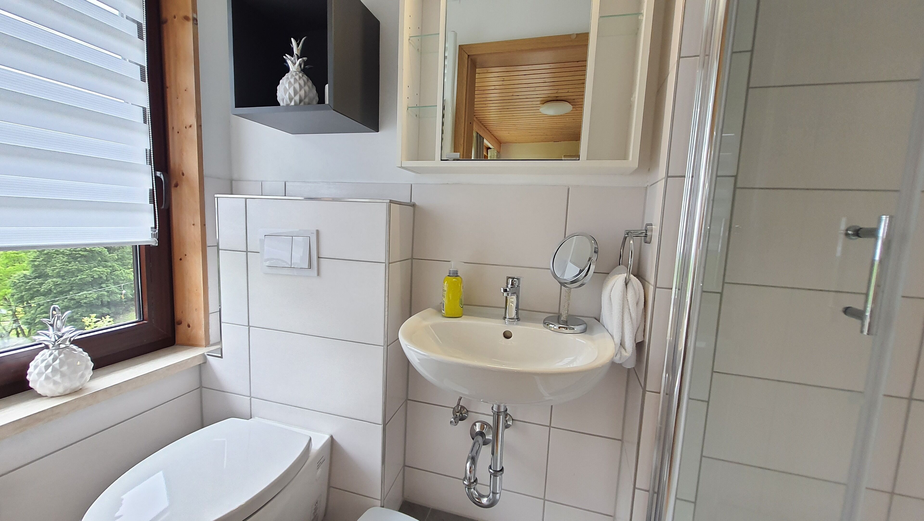 Un baño con inodoro, lavabo y ducha. Hay una ventana con persianas, un estante con una piña y un espejo sobre el lavabo.