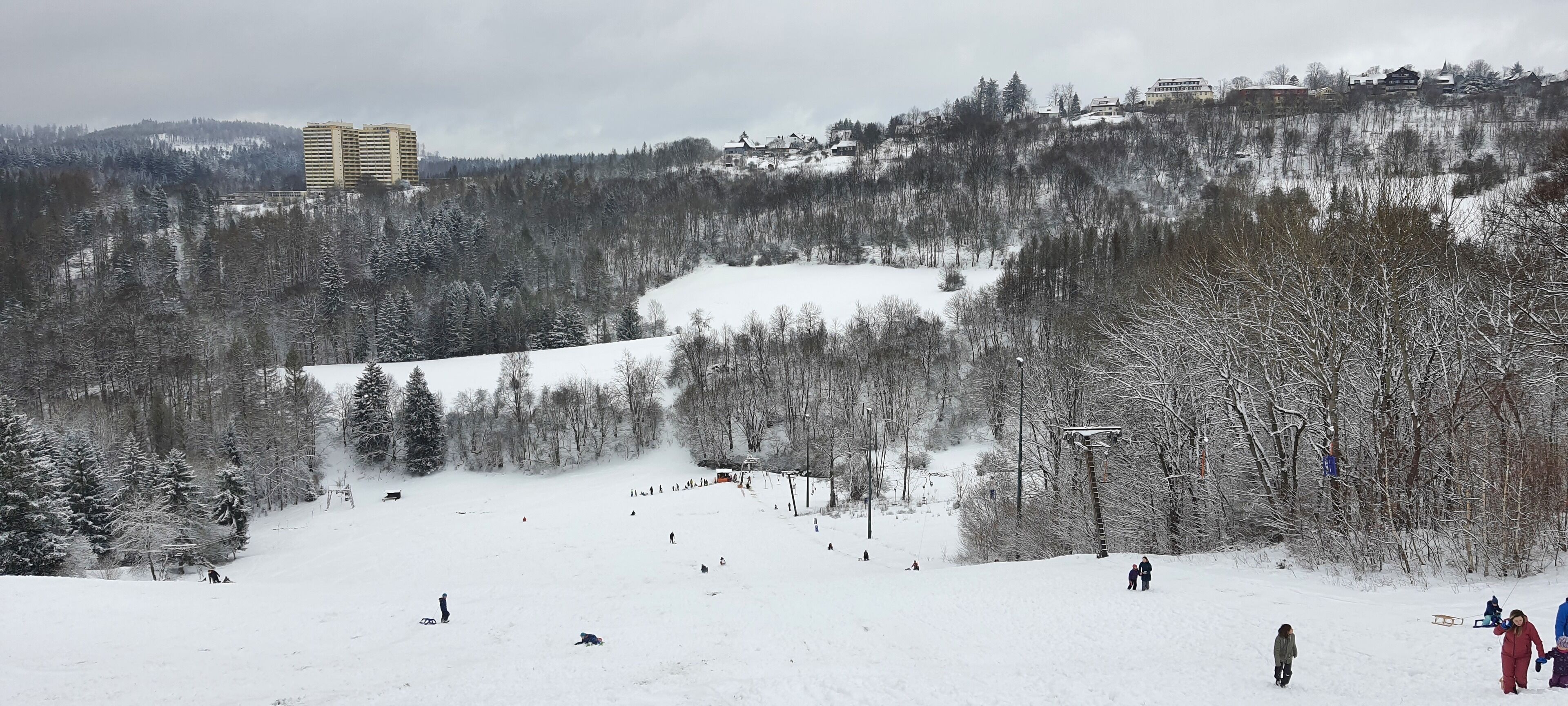A snowy ski slope with a ski lift, trees, and buildings in the background.