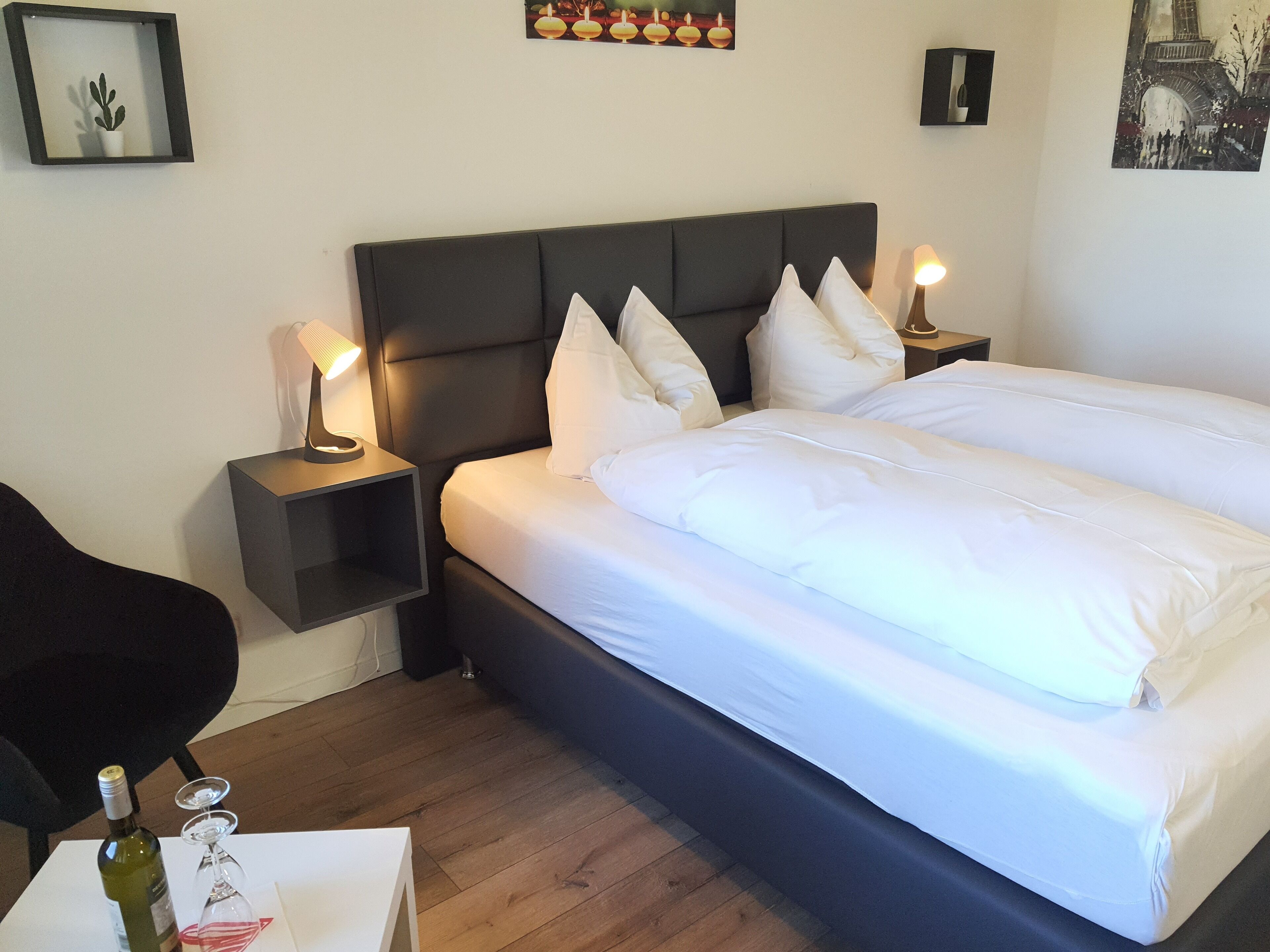 A neatly made bed with white linens and a dark headboard, flanked by two bedside tables with lamps, a small potted plant, and a bottle of wine on a side table.