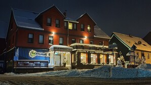 A snow-covered street with a row of buildings, including a brightly lit restaurant with outdoor seating. - Hotel Schneiderhof (Braunlage)