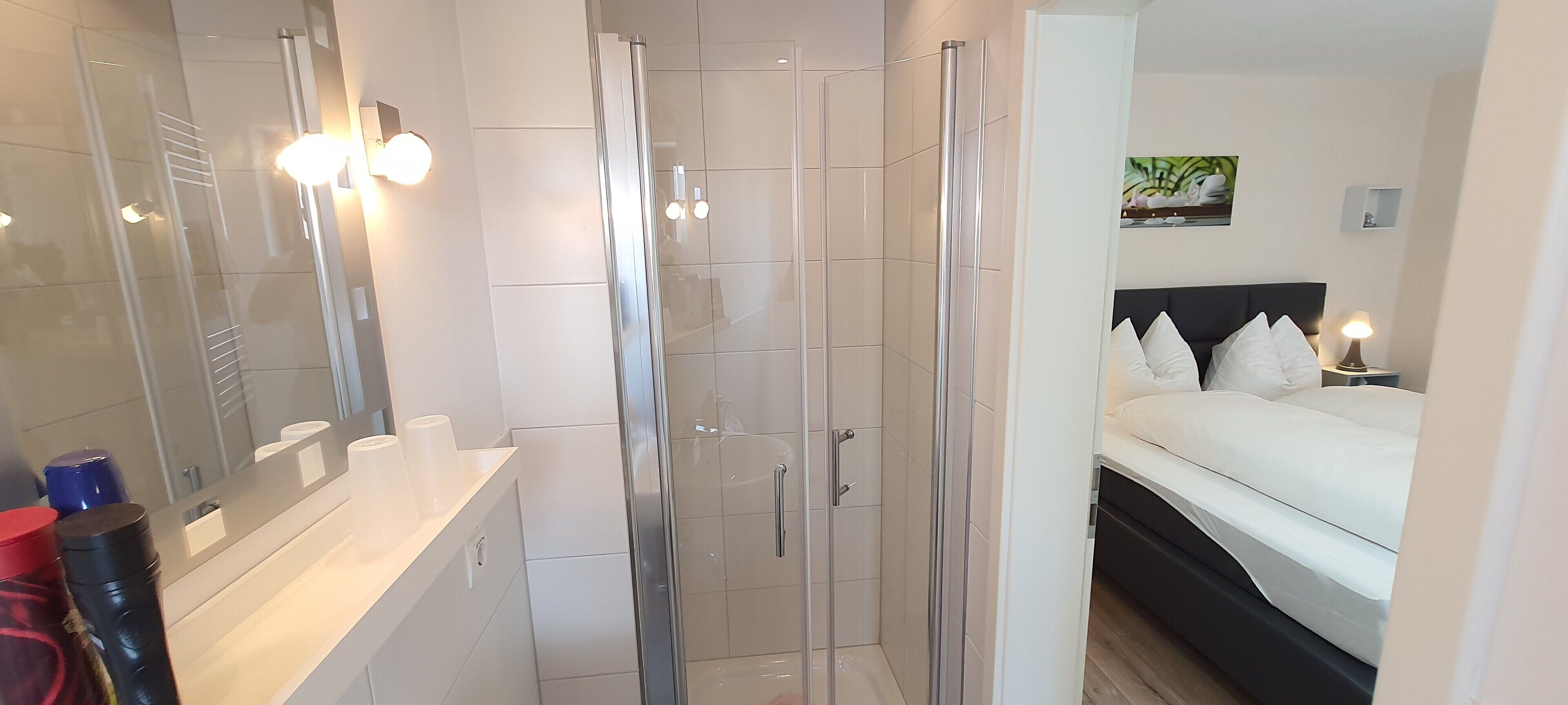 Een moderne badkamer met een glazen douchecabine, een wastafel met spiegel, en een bed met wit beddengoed in de aangrenzende kamer.