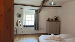 Een slaapkamer met een houten vloer, een bed met een witte deken, een dressoir, een raam met gordijnen en een hanglamp.