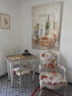 A room with a white table, a floral armchair, and a large painting of a sofa and fountain. - Charming old country house ( 70m2 ) for 2 to 4 persons (Brue-Auriac)