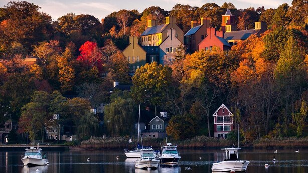 A lakeside view with boats docked, surrounded by autumn-colored trees and houses. - Captain's Cottage - Cold Spring Harbor Water Views (Cold Spring Harbor)