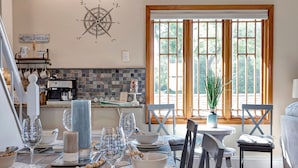 A dining area with a table set for four, a coffee machine, and a window with a view of trees. - Captain's Cottage - Cold Spring Harbor Water Views (Cold Spring Harbor)