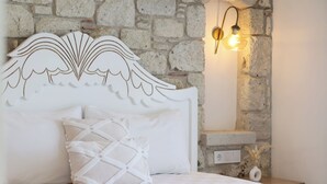 Un lit avec une tête de lit blanche, un coussin décoratif, une table de chevet avec une lampe fixée au mur et un mur en pierre.