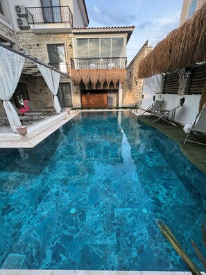 Une piscine à l’eau d’un bleu cristallin, entourée de chaises longues et d’un bâtiment en pierre doté d’un balcon.