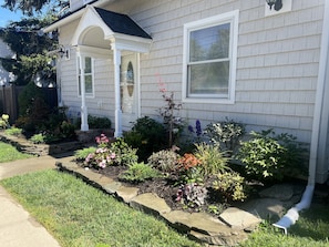 Una casa con porche cubierto, un jardín con diversas plantas y flores, y un bajante visible.