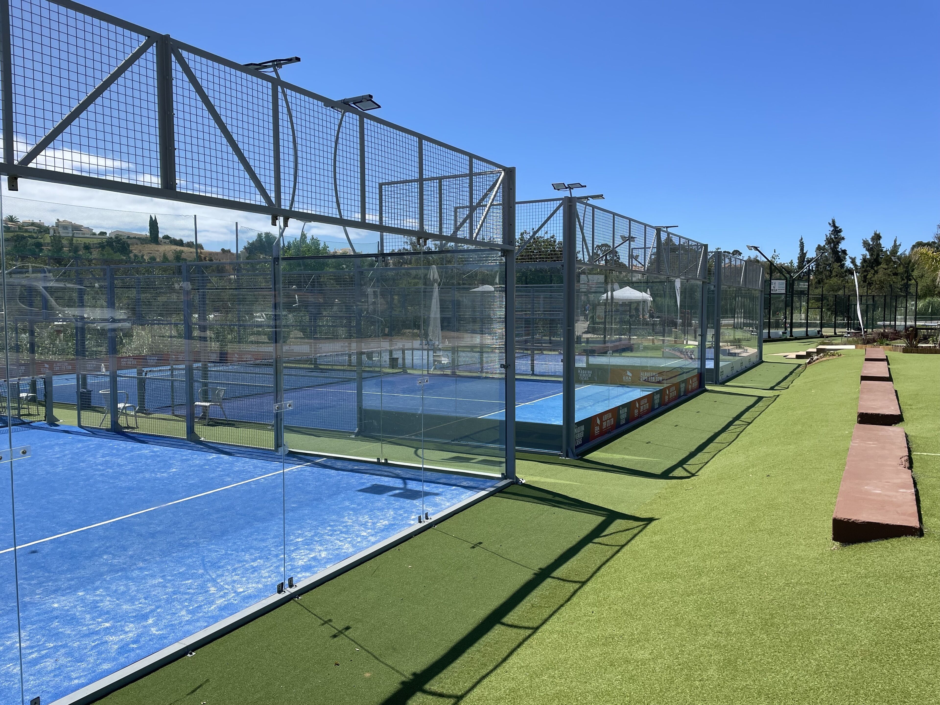 Een sportcomplex met meerdere velden, een groen kunstgrasgedeelte en een helderblauwe lucht.