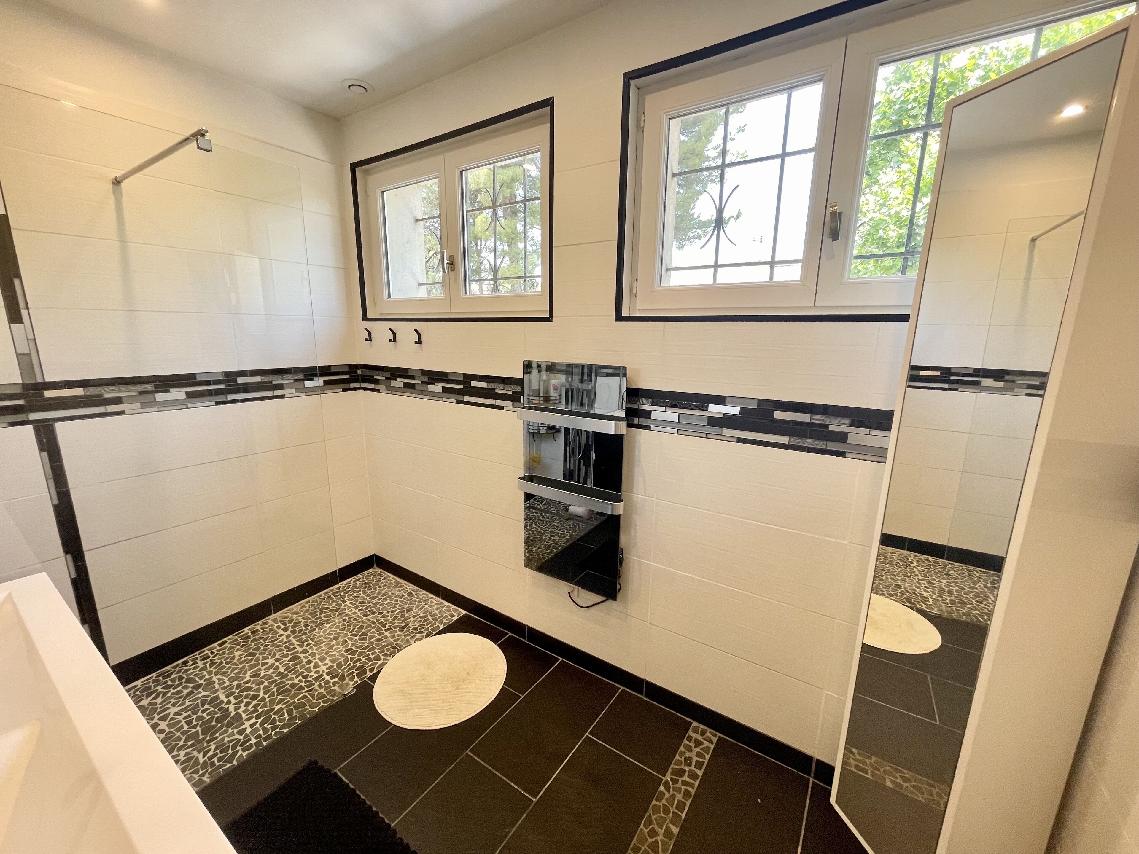A modern bathroom with white tiles, a black and white patterned border, a built-in sink, a mirror, and a window with a view of trees.