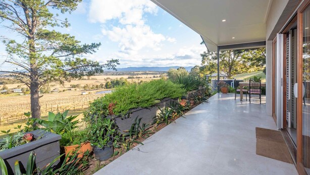A balcony with a view of a vineyard, a stone wall with plants, and a glass door leading to a patio area.