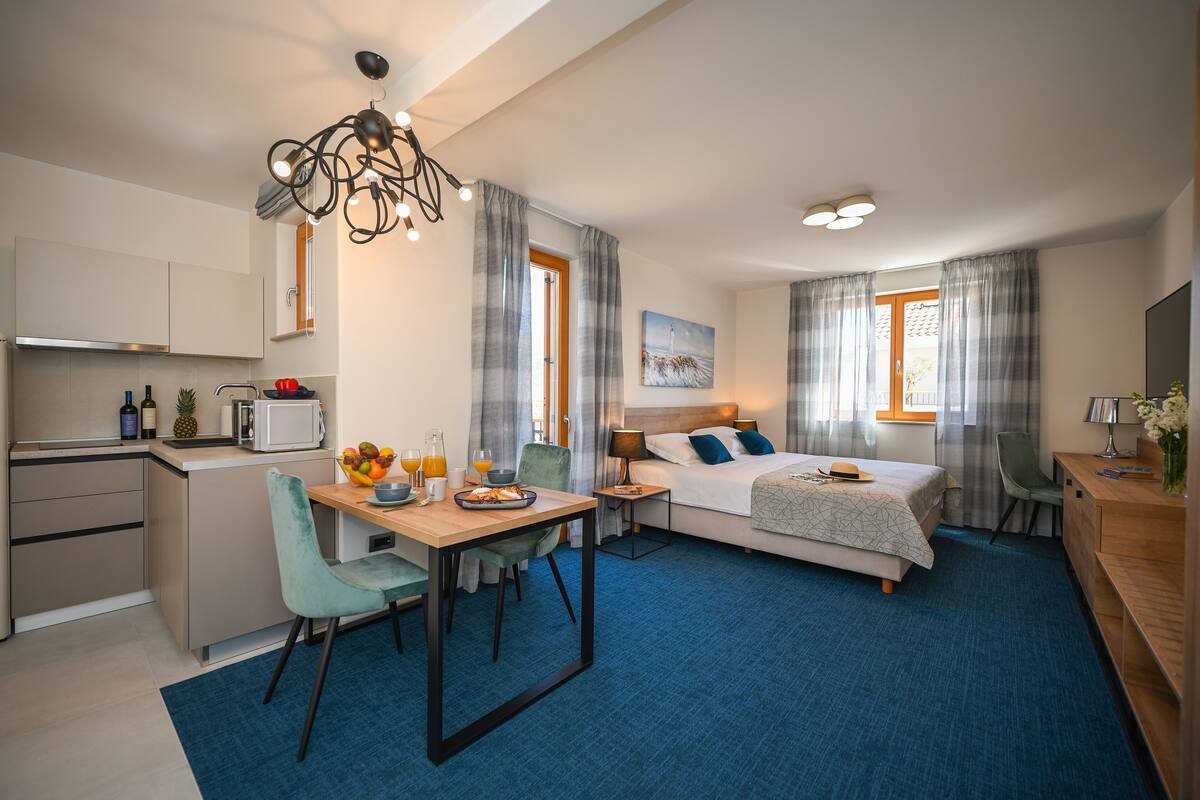 A modern hotel room with a kitchenette, a dining area, a bed with a view, and a desk with a lamp.