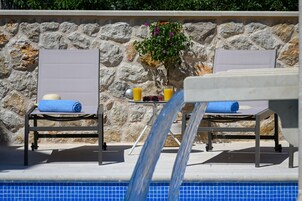 Zona junto a la piscina con dos sillas reclinables, una mesa con bebidas y un muro de piedra.