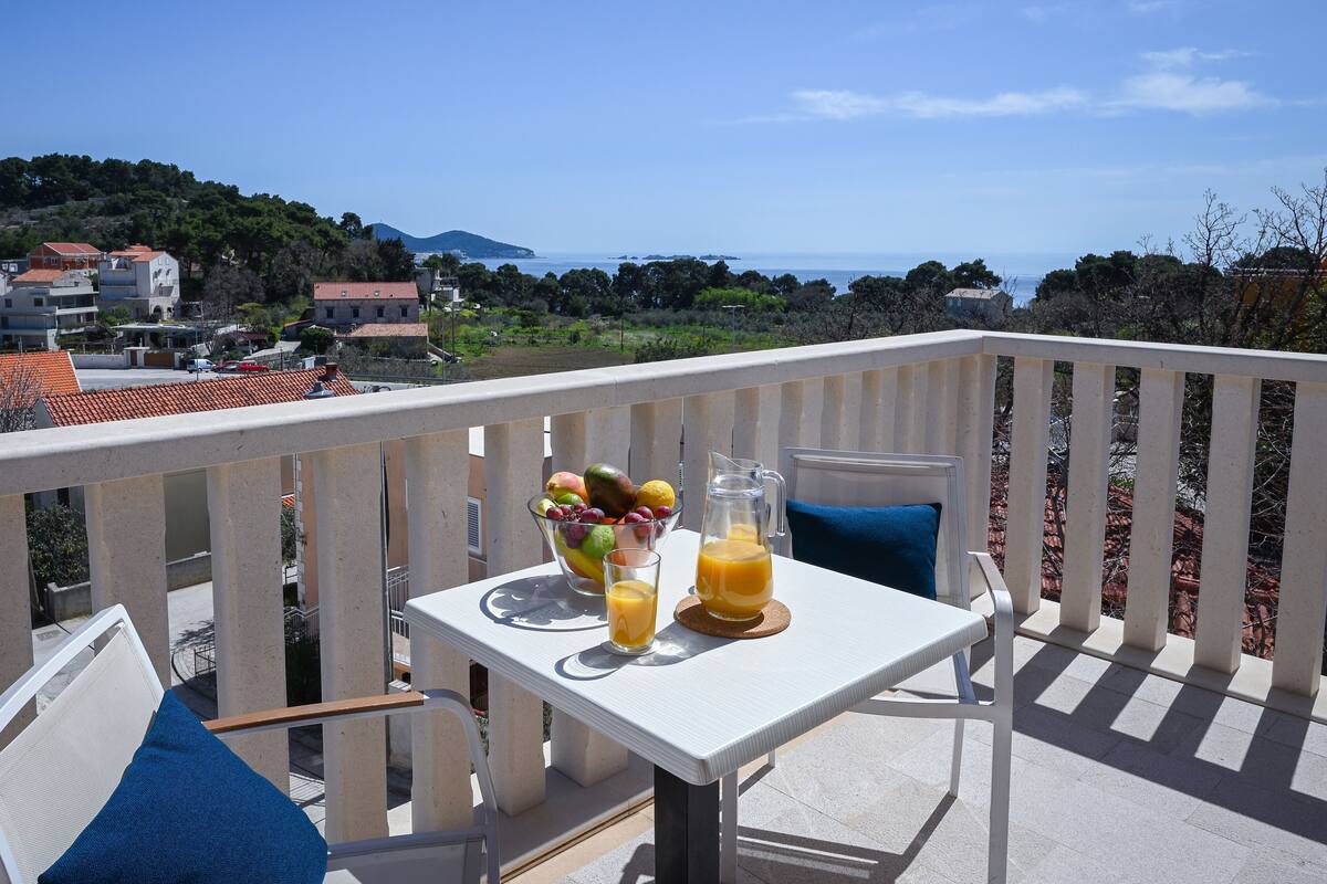 A balcony with a table set for breakfast, featuring a fruit bowl, a glass of orange juice, and a pitcher of orange juice.